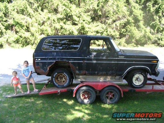 Side view of a rusty black 1978 Ford Bronco on a trailer, with two small children standing on a ramp next to it