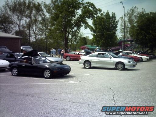 Several convertibles and coupes parked in a lot, some with hoods up. Trees and a building in background