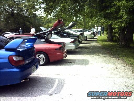 Several cars parked in a line next to trees, some with hoods or trunks open. A blue Subaru WRX is in the foreground