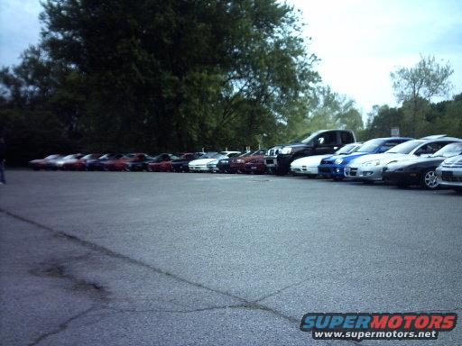 Several cars parked in a row, including a black truck and various sports cars. A Supermotors logo is visible at the bottom right
