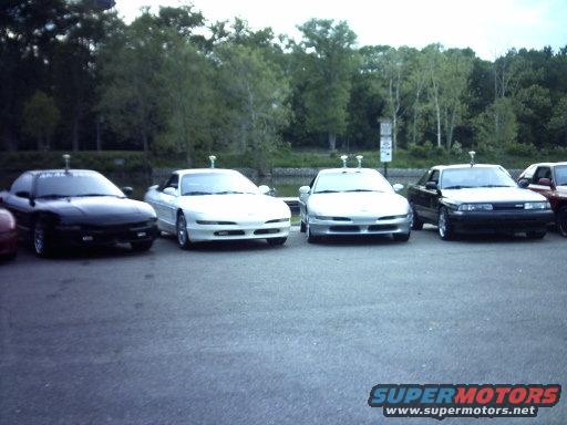 Four coupes parked in a lot, three are silver or white and one is black. The front of the cars are visible