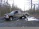 2002 Ford F-350 - A dark 2002 Ford F-350 pickup truck with dual rear wheels navigates a muddy and snowy dirt road through a wooded area.