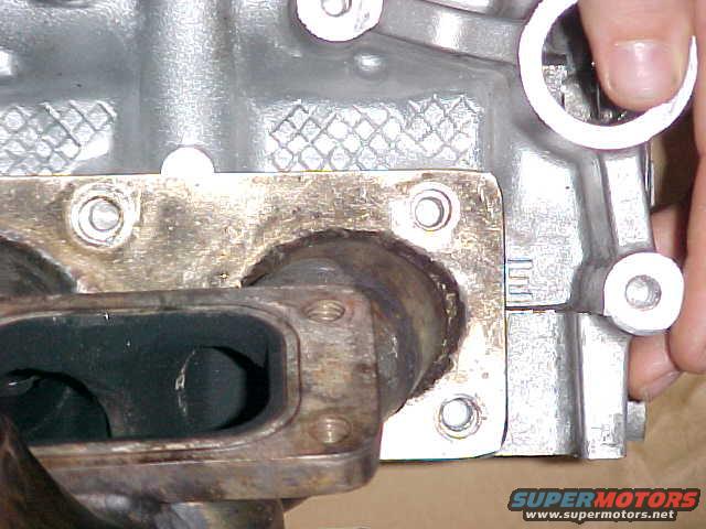 Close-up of an exhaust manifold bolted to a silver engine block. A hand is visible on the right