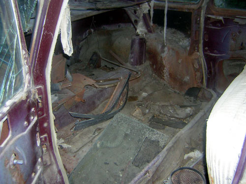 Interior of a very run-down car, showing rusted floor, torn seats, and debris