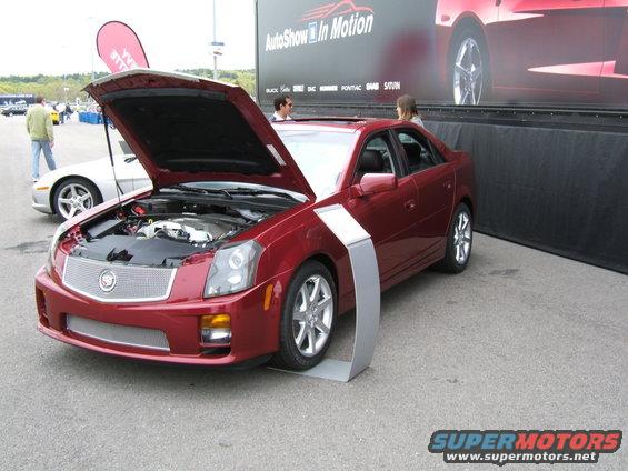 Red Cadillac sedan, hood open, showing engine. Silver trim piece in front of front wheel. Other cars and a banner in background