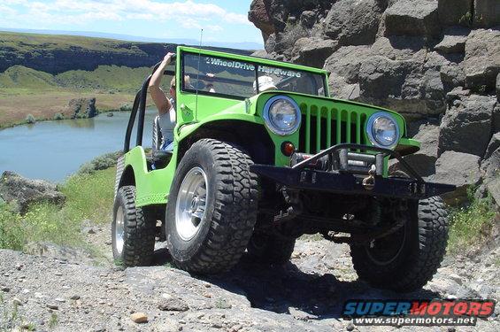 Green Jeep CJ with off-road tires driving on a rocky trail near a body of water. Text on windshield reads '4WheelDrive Hardware'