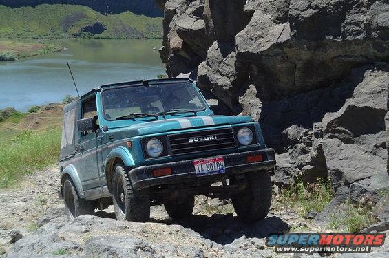 Teal Suzuki Samurai driving on a rocky trail next to a cliff and a body of water