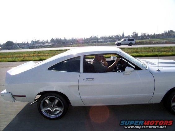 White 1975 Ford Mustang II coupe with a man driving, seen from the side on a highway