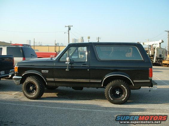Black 1989 Ford Bronco parked outside, side view. Other vehicles and buildings in background