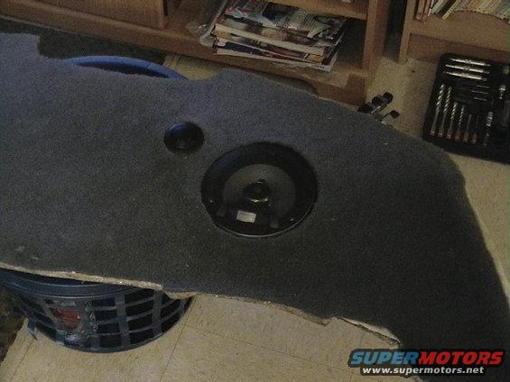 Carpeted panel with a speaker cutout and a separate small black circular object, resting on a black laundry basket