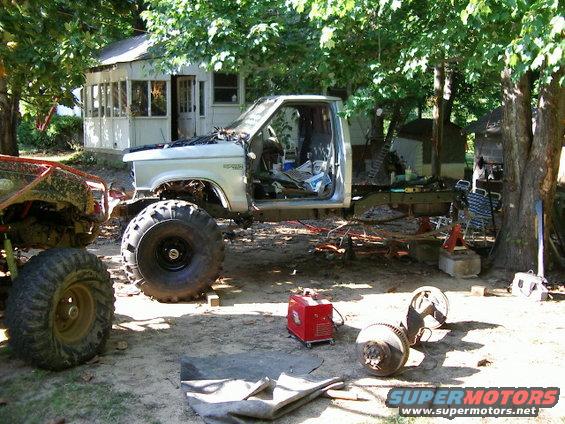 Pickup truck chassis with large tires, open doors, and hood. Another off-road vehicle is to the left. Parts and a welder are on the ground