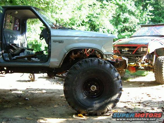 Ford Ranger pickup truck with large tires, missing doors and hood. Another off-road vehicle is parked next to it