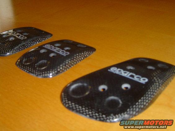 Three black Sparco pedal covers with carbon fiber pattern on a wooden surface