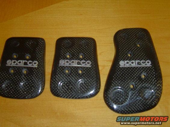 three carbon fiber Sparco pedal covers on a wooden surface