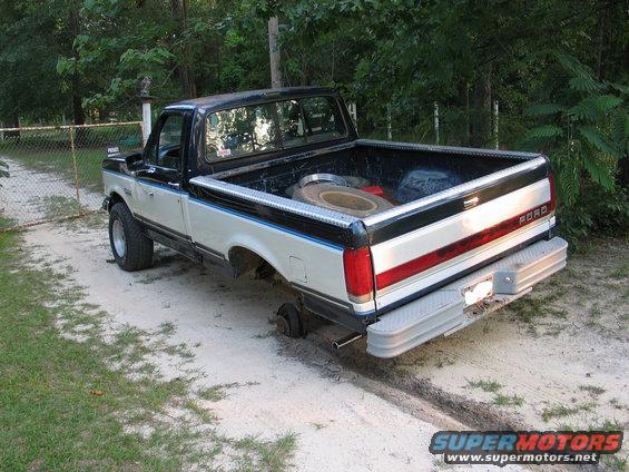 Two-tone Ford pickup truck missing rear passenger-side wheel, with spare tire in bed
