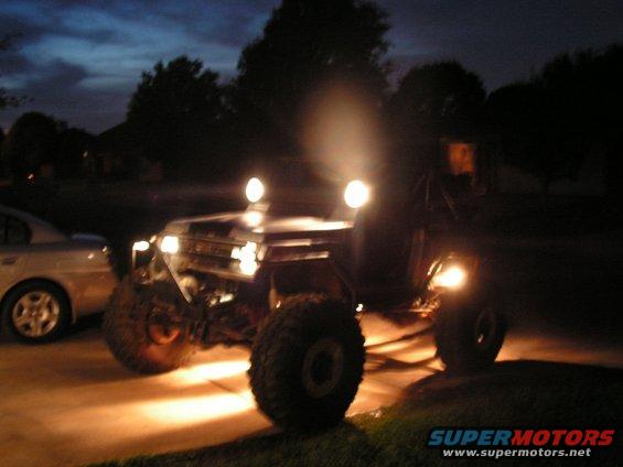 Off-road SUV with big tires and headlights on, parked next to a sedan at dusk