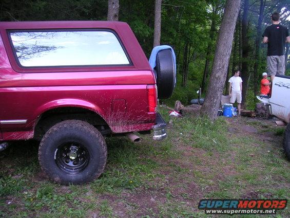 Red SUV with spare tire on back, parked in a wooded area. Another white truck is nearby with people standing around a campfire
