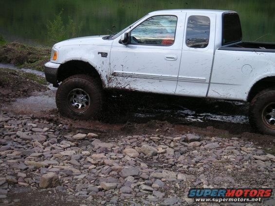 White pickup truck with mud splattered on its side, driving over rocks and through water