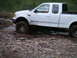 1994 Ford Bronco - A white pickup truck navigates a rocky, muddy terrain near a body of water.