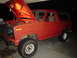 1981 Ford Bronco - The front end of a red SUV is visible with the hood open, revealing the engine bay under a work light.