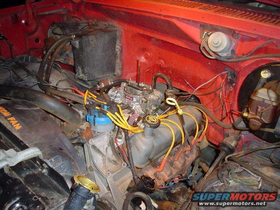 Engine bay of a red 1981 Ford Bronco, showing the V8 engine, carburetor, distributor, and battery