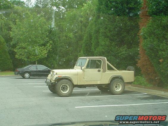 Tan Jeep CJ-7 parked in a lot, side view. A black sedan is parked behind it