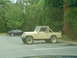 1981 Ford Bronco - A tan Jeep CJ-7 is parked in a lot next to a black sedan, with trees in the background.
