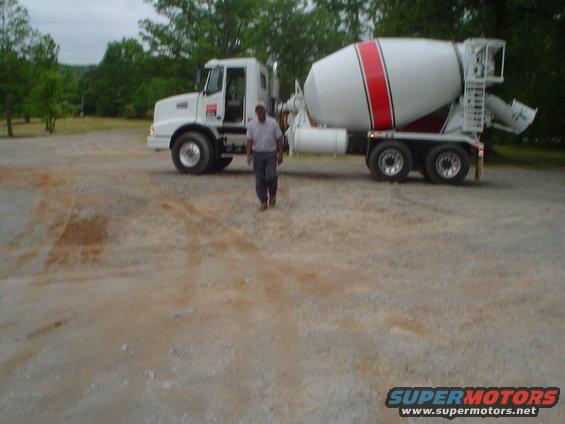 Cement mixer truck with a man standing in front of it on a gravel lot