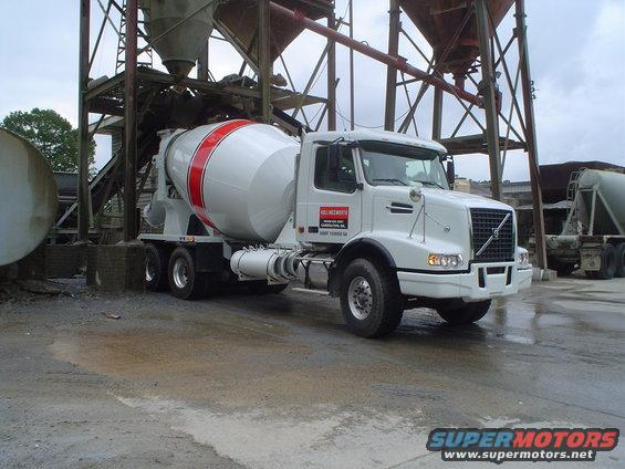 White concrete mixer truck with red stripes on the drum, parked on wet ground near industrial silos