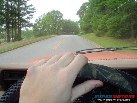 Hand on steering wheel of a vehicle driving down a two-lane road with trees and grass on the sides