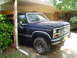 1981 Ford Bronco - Dark blue Ford Bronco parked under a carport, front quarter view. The hood is slightly ajar.