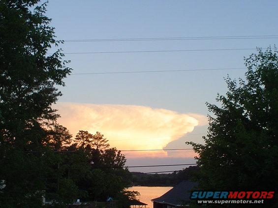 Sky with a large, illuminated cloud formation over a body of water, framed by trees