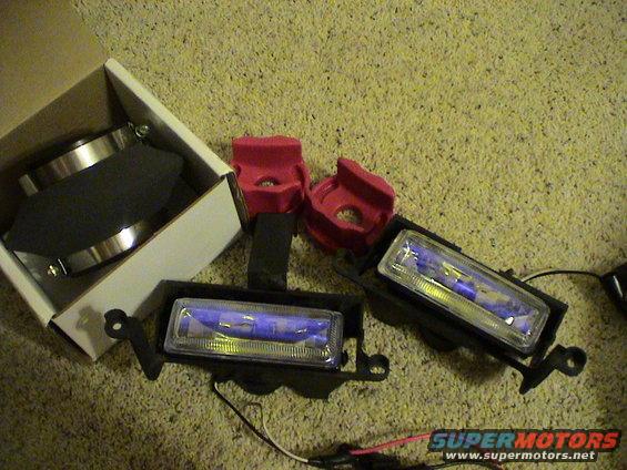 Two fog lights and parts of an air intake kit are on a carpeted surface. One fog light is in its box