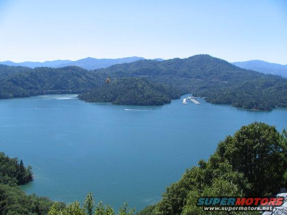 View of a large blue lake surrounded by tree-covered mountains under a clear sky. A small boat is visible on the water