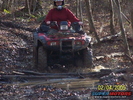 Red ATV splashing through mud and water on a wooded trail