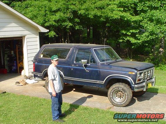 Dark blue Ford Bronco parked in a driveway next to a garage, person standing beside it