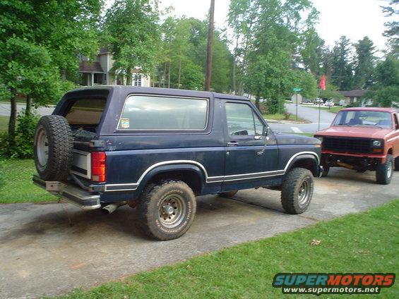 Dark blue 1981 Ford Bronco with spare tire on rear, parked next to a red pickup truck