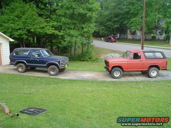 Two Ford Broncos parked on a driveway, one blue and one orange. Trees and houses in background