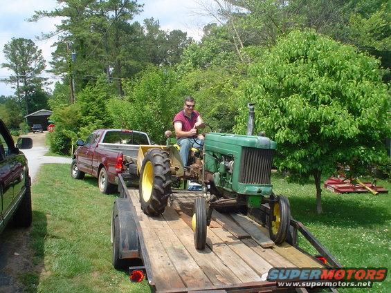 Green tractor on a wooden trailer being towed by a red pickup truck