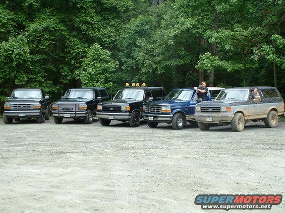Five Ford Bronco SUVs and trucks parked in a gravel lot with trees in the background