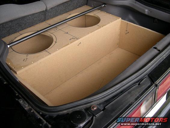 Custom MDF subwoofer box with two circular cutouts and drawn measurements inside a car trunk