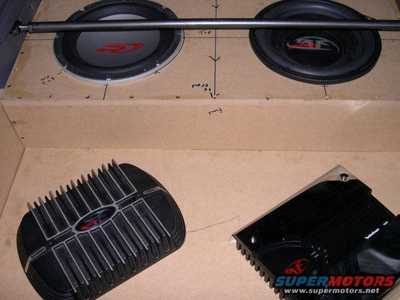 Two car subwoofers mounted in a wooden enclosure, with two car amplifiers below. Markings on enclosure indicate measurements