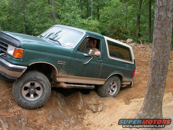 Green 1990 Ford Bronco navigating rocky terrain off-road. Driver visible