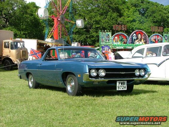 Blue 1970 Ford Ranchero parked on grass with a Ferris wheel and carnival rides in the background