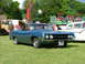 1970 Ford Ranchero - A blue 1970 Ford Ranchero is seen from the front, parked on a grassy field with carnival attractions behind it.