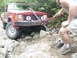 1985 Ford F-150 - A person uses a hi-lift jack to free a red Jeep Comanche pickup truck from a rocky obstacle on a trail.