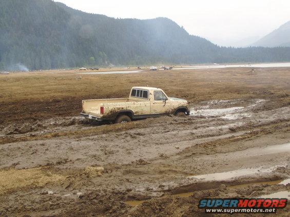 Beige pickup truck stuck in deep mud, front wheels submerged. Other vehicles visible in the distance