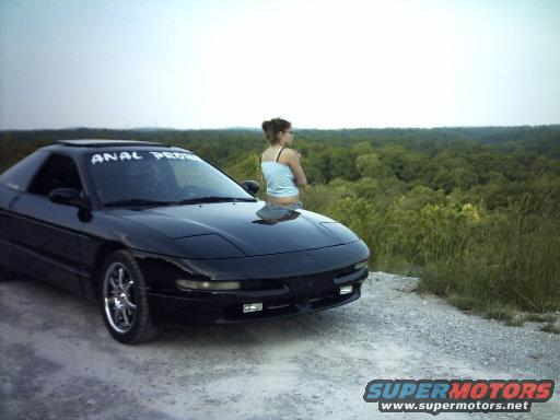 Black Ford Probe parked on a dirt road with a woman on the hood, forest in the background