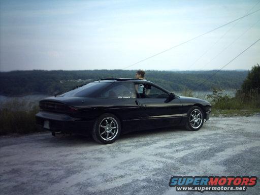 Black 1995 Ford Probe parked on a gravel road with a person in the driver's seat. Overlooks a body of water and forested hills