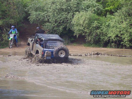 Off-road vehicle splashing through mud, dirt bike nearby